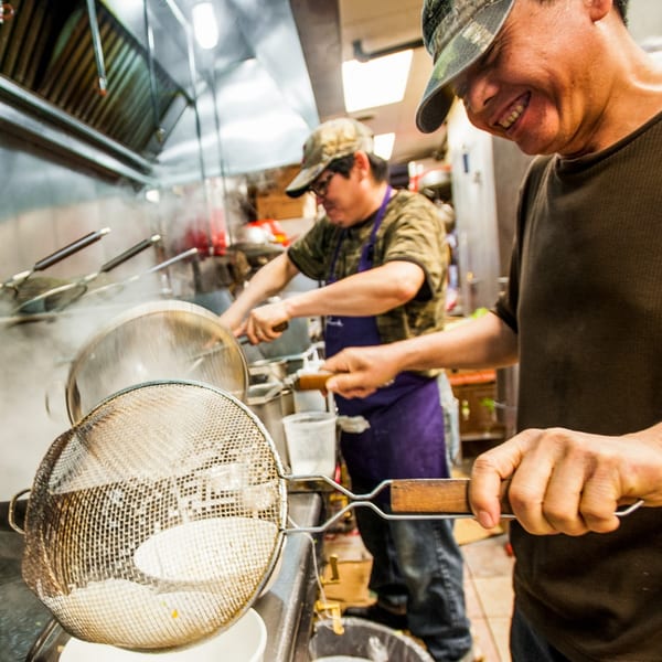 Two Vietnamese chefs in a steamy kitchen, one smiling while straining noodles.