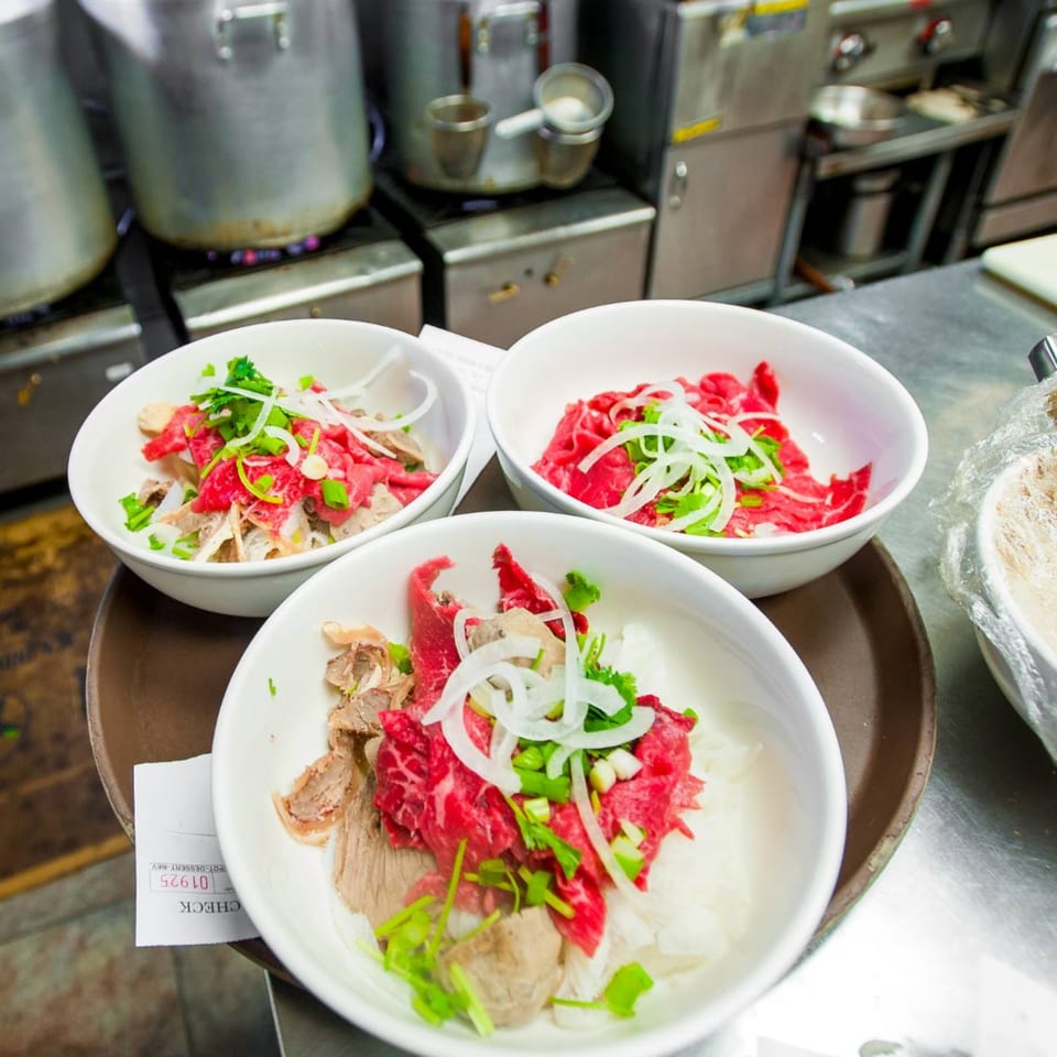 Three bowls of Tank Noodle's Vietnamese pho with various meats, herbs, and onions in a kitchen setting.