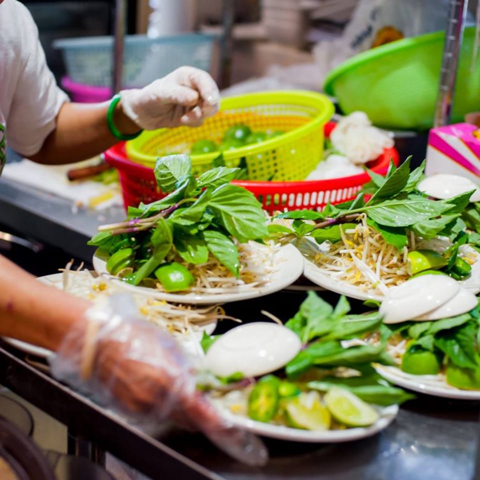 A gloved hand prepares plates of fresh Vietnamese herbs, bean sprouts, and lime wedges.
