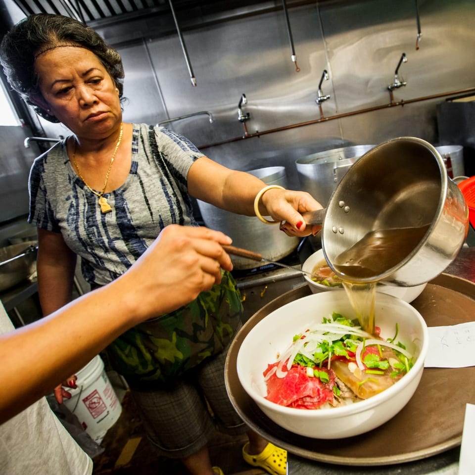 A Tank Noodle chef pours broth into a bowl of pho with meat, onions, and herbs.