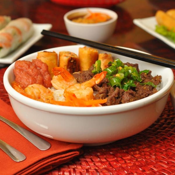 A Vietnamese noodle bowl with shrimp, beef, spring rolls, and a red sauce, with chopsticks on top.