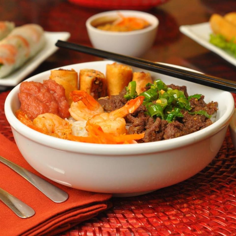 A Vietnamese noodle bowl with shrimp, beef, spring rolls, and a red sauce, with chopsticks on top.