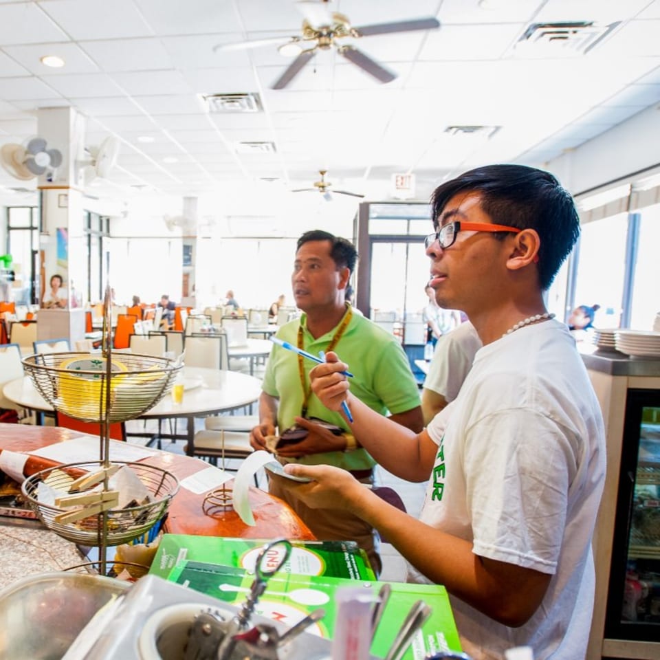 Two men stand at a counter in a brightly lit Vietnamese restaurant, one holding a pen.